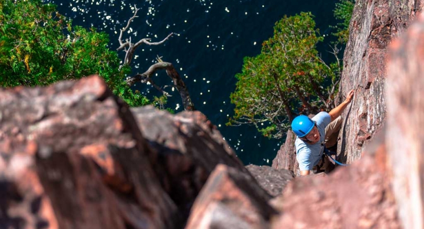 A person wearing safety gear and secured by ropes climbs a rock wall high above a lake. 